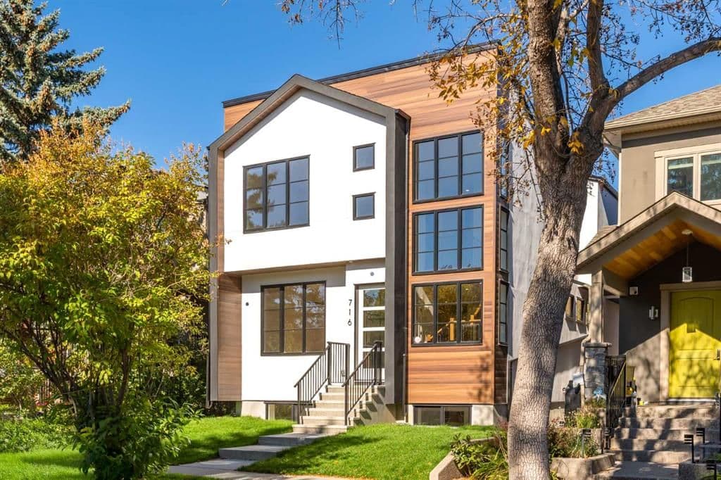 A newly built two-storey infill home in a Calgary neighbourhood, white stucco and wood siding with black-framed windows, on a landscaped lot lined with autumn trees.
