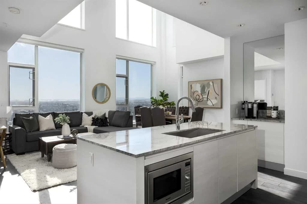 A bright, staged open-concept Calgary condo, a marble-topped kitchen island opening onto a living room with floor-to-ceiling windows and a city view.